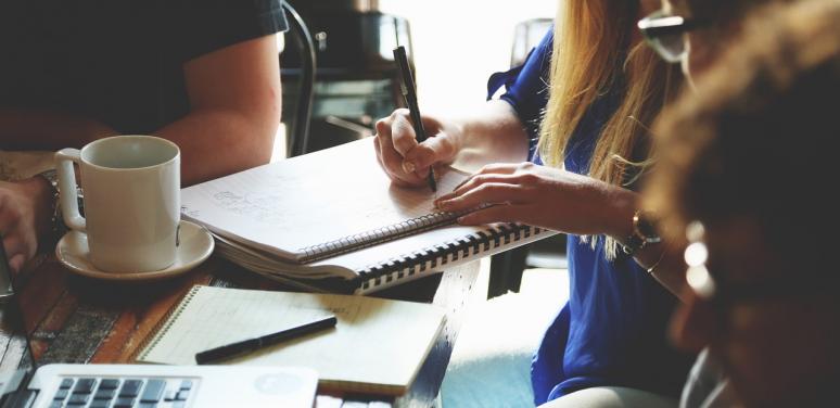 people at a desk working, with focus on the desk and someone writing on a notebook