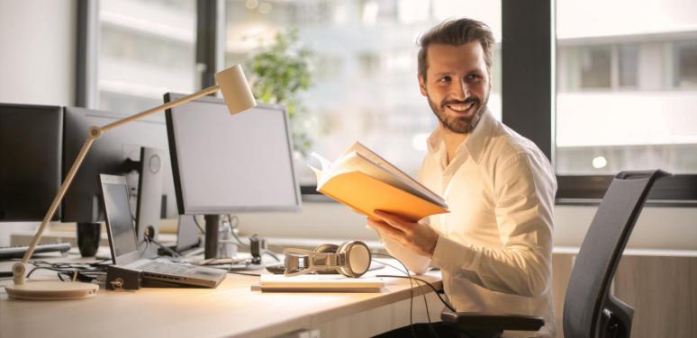 a man with a book smiles at a computer screen while glancing off to the side.
